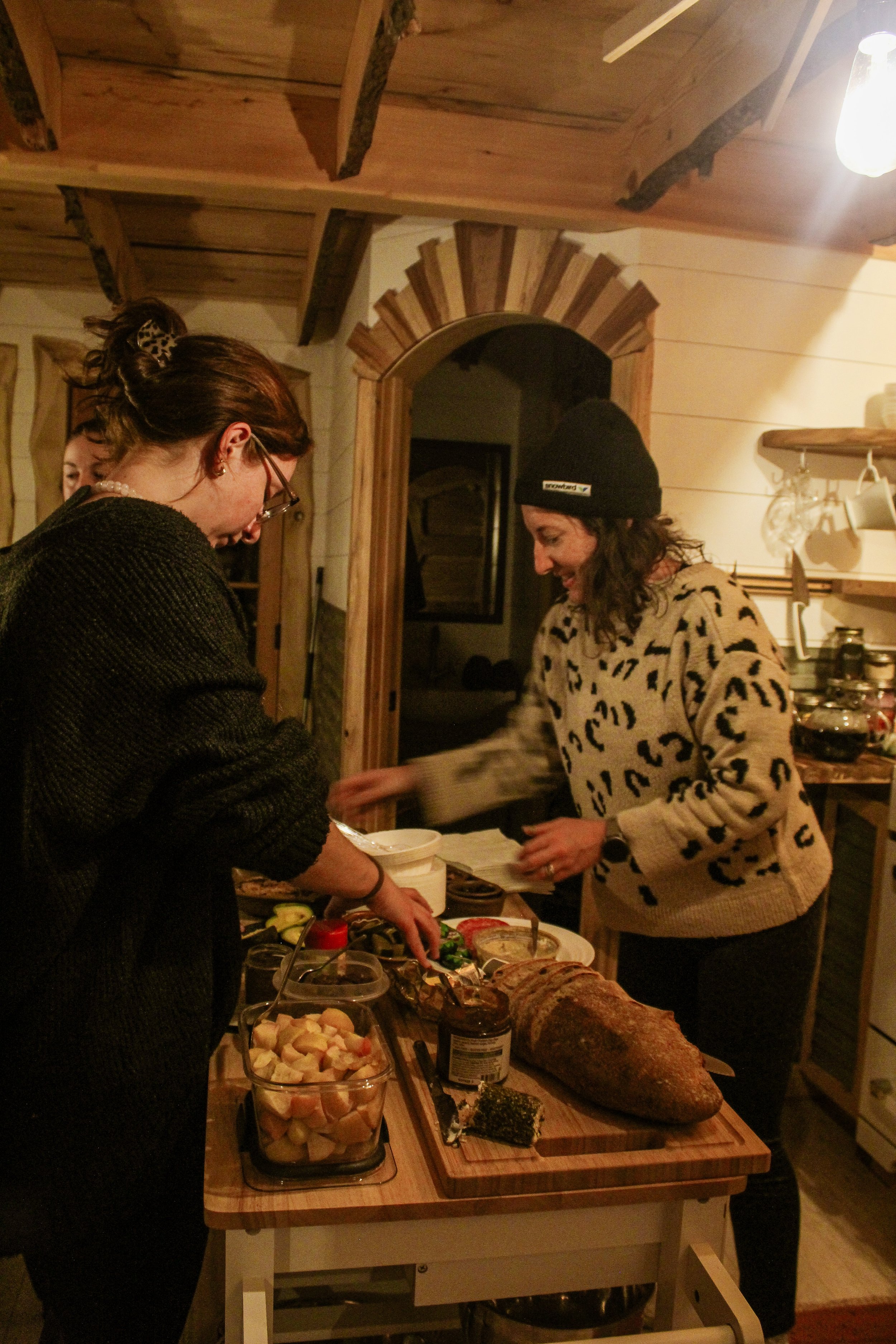 Two women are preparing food at a rustic wooden kitchen counter with various ingredients and a loaf of bread on it. They are smiling and engaging in conversation.