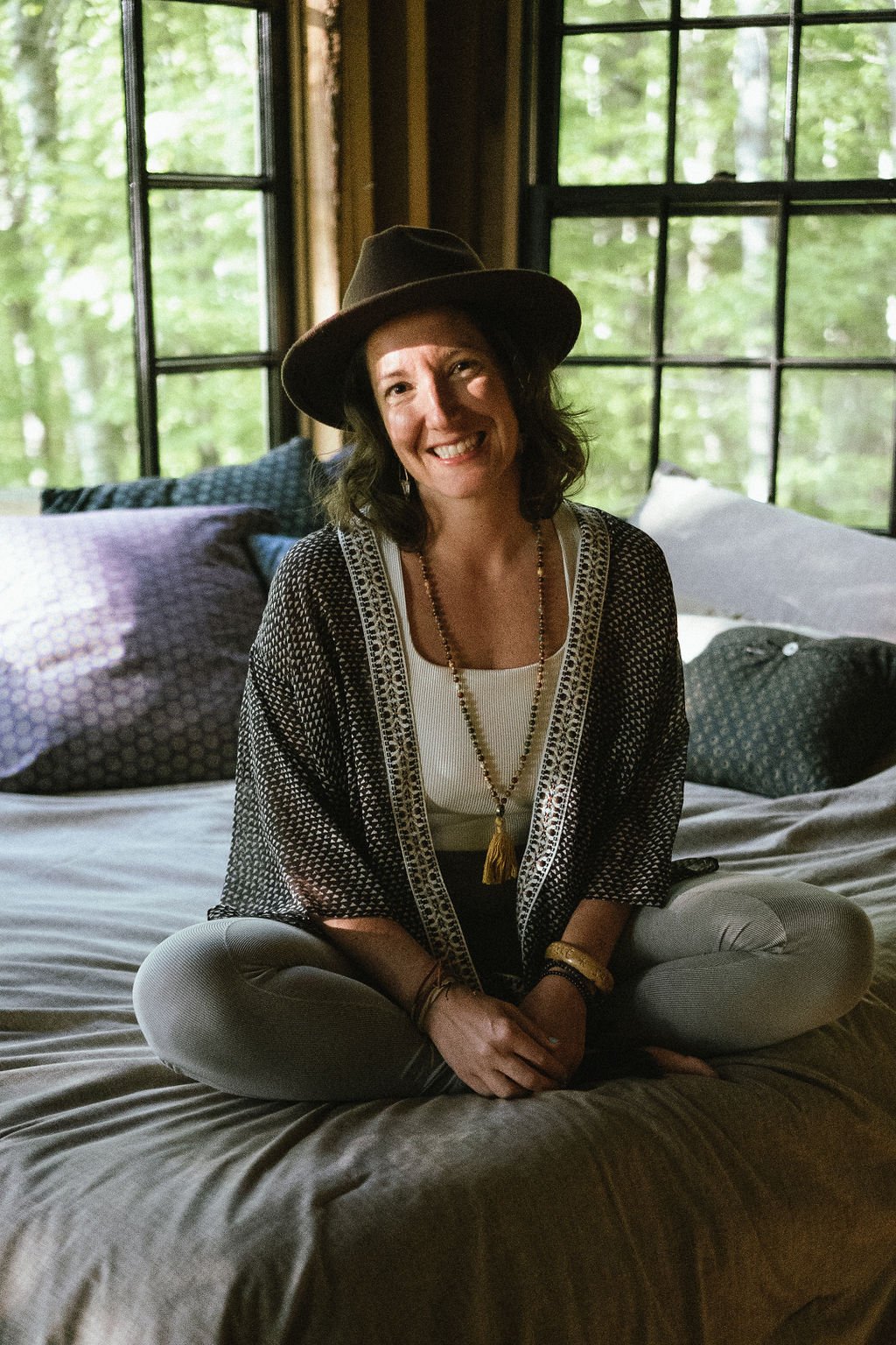 A smiling woman sitting cross-legged on a bed, wearing a brown hat, a white top, a patterned open cardigan, and jewelry, with large windows showing green trees outside in the background.