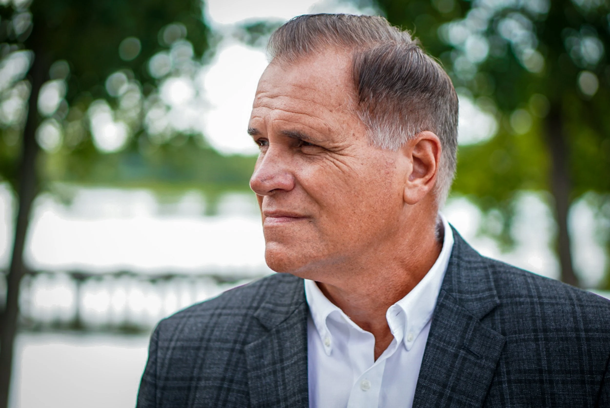 Profile of a middle-aged man with short gray hair and facial wrinkles, wearing a checkered blazer and white shirt, outdoors with blurred trees and water in the background.