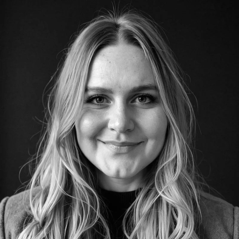 Black and white portrait of a smiling young woman with long wavy hair, wearing a dark top, against a dark background.