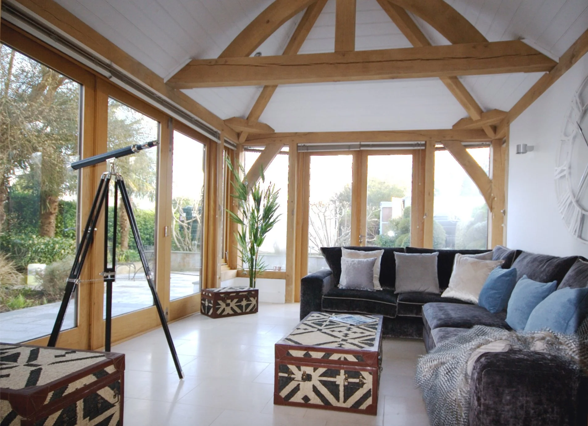 A rich chocolate brown sofa has cream and blue cushions and a feathered throw.  There are aztec style coffee and side tables and a telescope stands in the french window