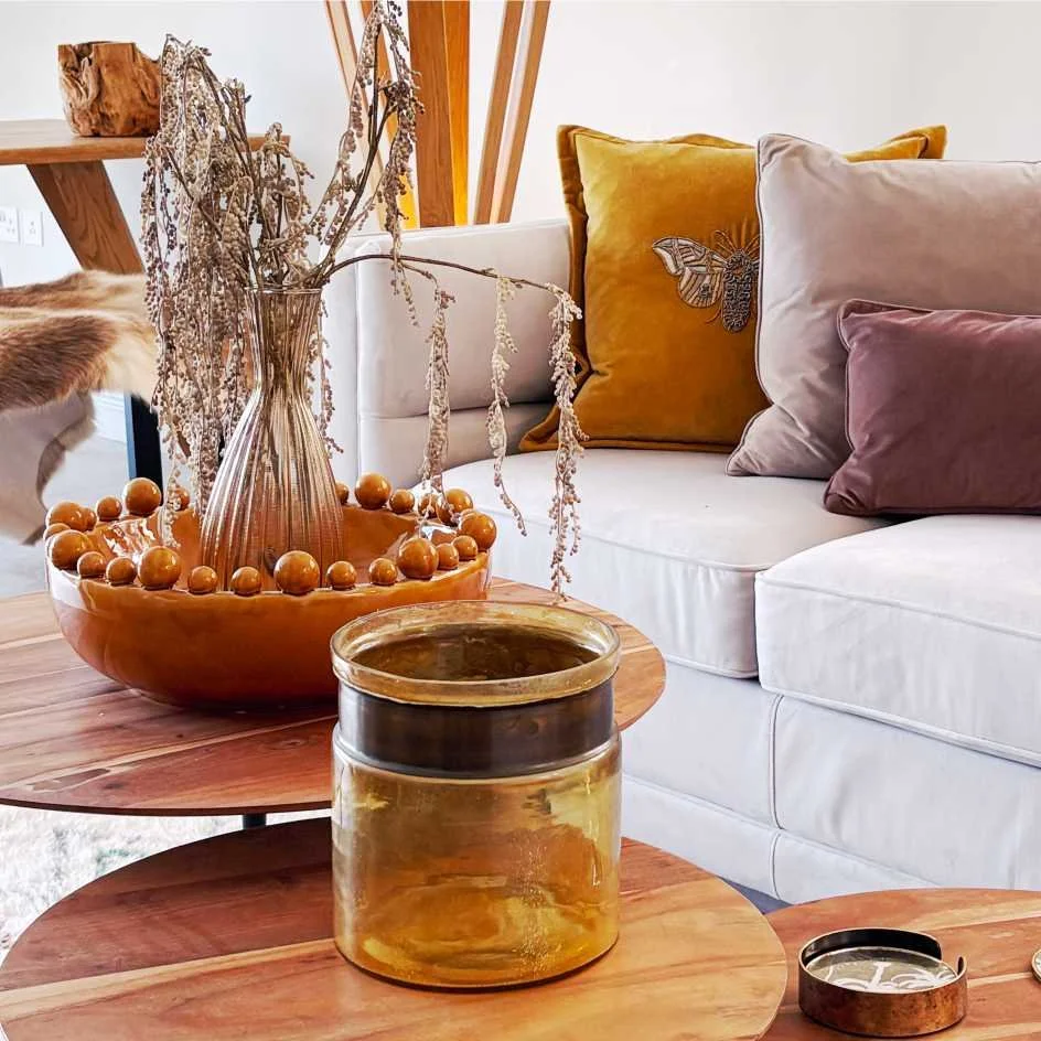 A laid back lounge with natural wood, grasses and fur throws.  A comfy sofa has cushions in ochre and yellow and there is a ball rimmed bowl and glass yellow vase on the round nest of coffee tables