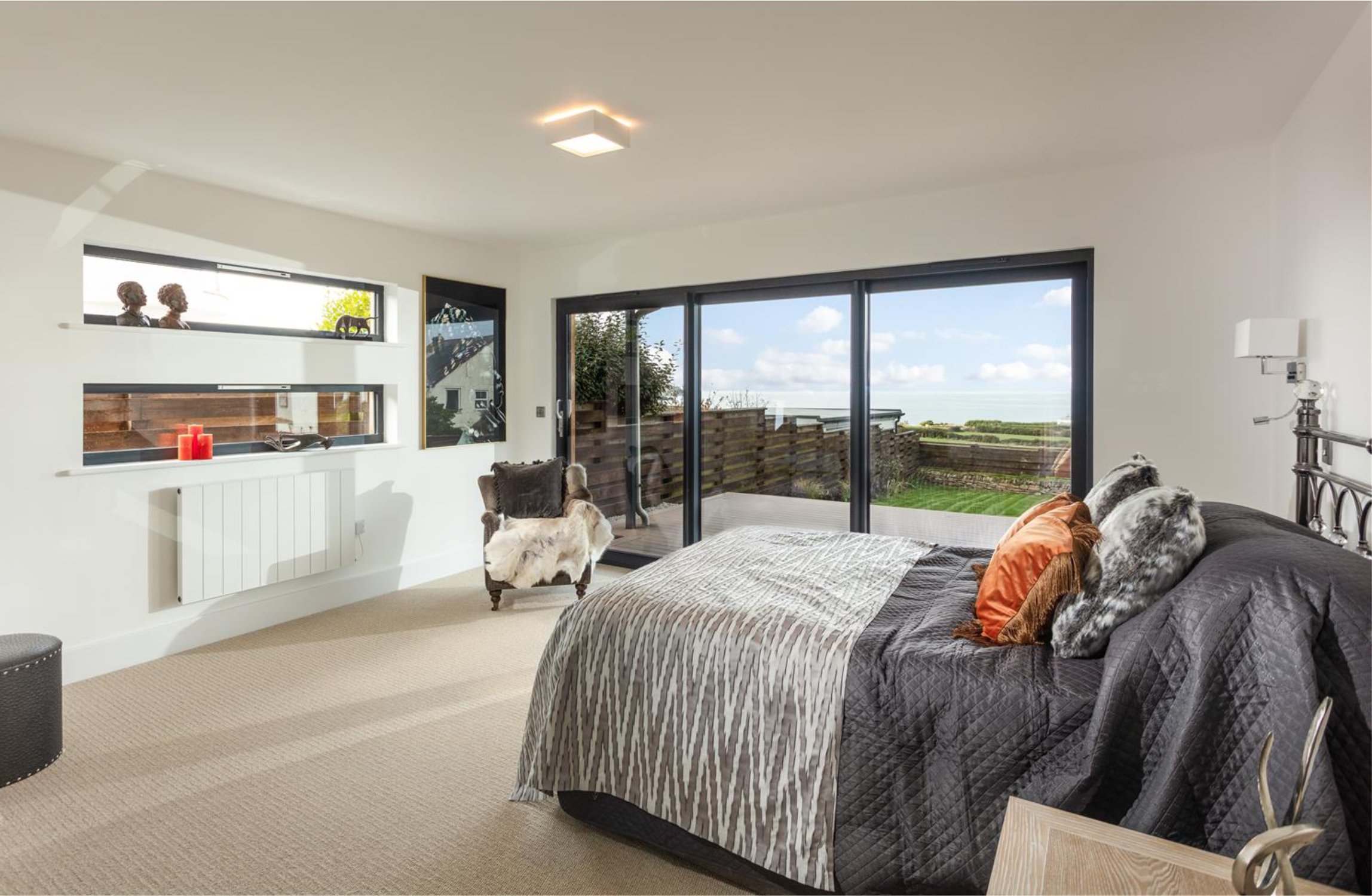 Architectural bedroom with black frame window and doors.  A bed with grey bedspreads and throw have orange cushions