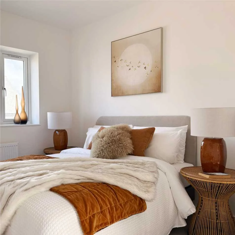 A beige picture of birds hangs above a bed that has a white bedspread and is layered with orange and brown throws and pillows.  Two lamps with russet ceramic bases are on wicker bedside tables and curvy brown glass vases decorate the window ledge