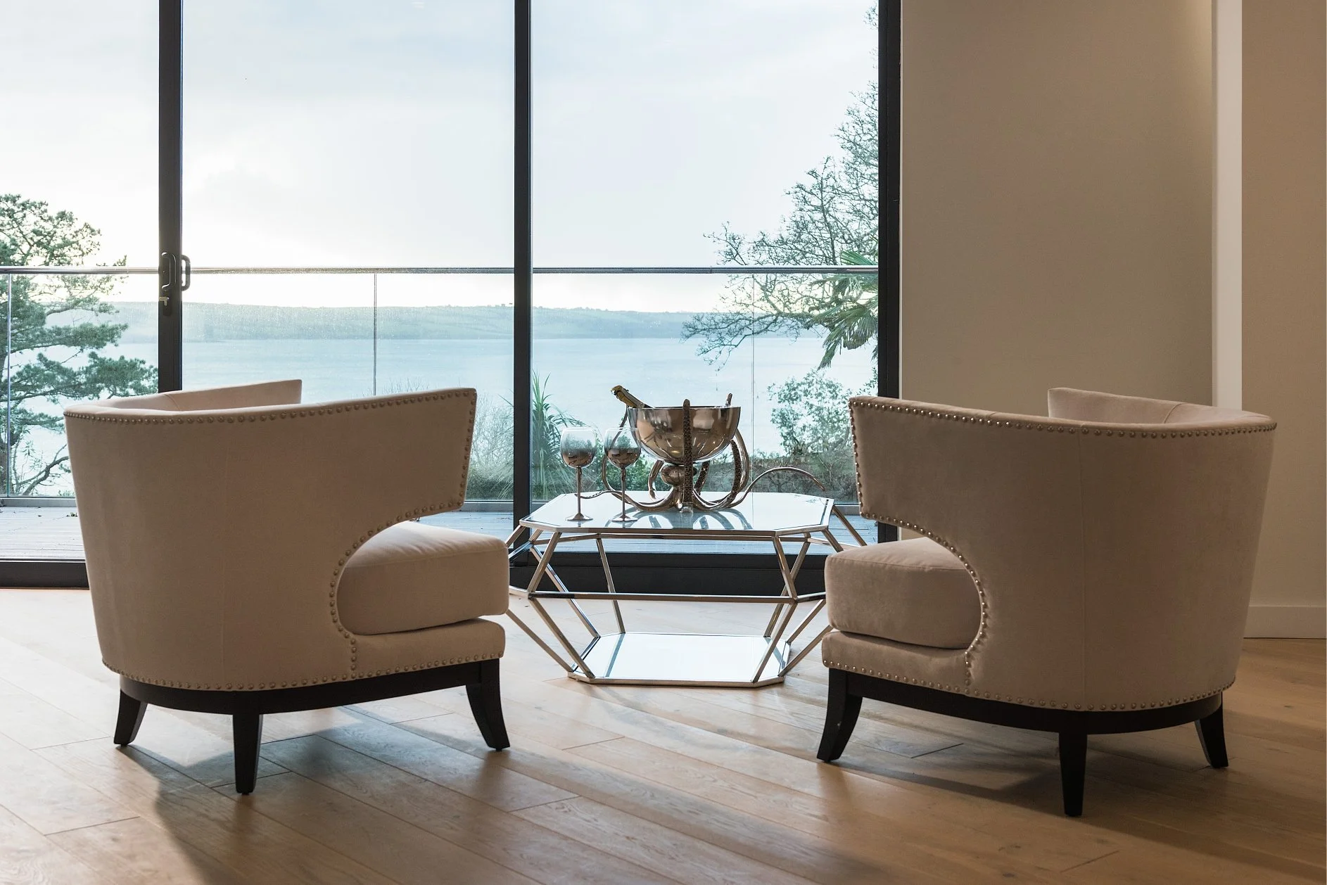two cream round back chairs with stud detailing and dark wood legs, chrome coffee table and octopus bowl with champagne and glasses.  All overlooking an estuary