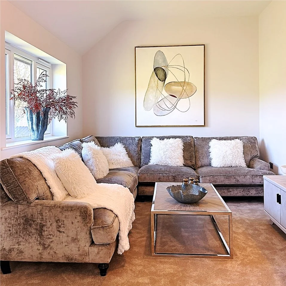 Large grey velvet corner sofa is decorated with white throws and fluffy cushions.  Autumnal leaf spray is in a blue wavy vase and abstract art ties all the colours in