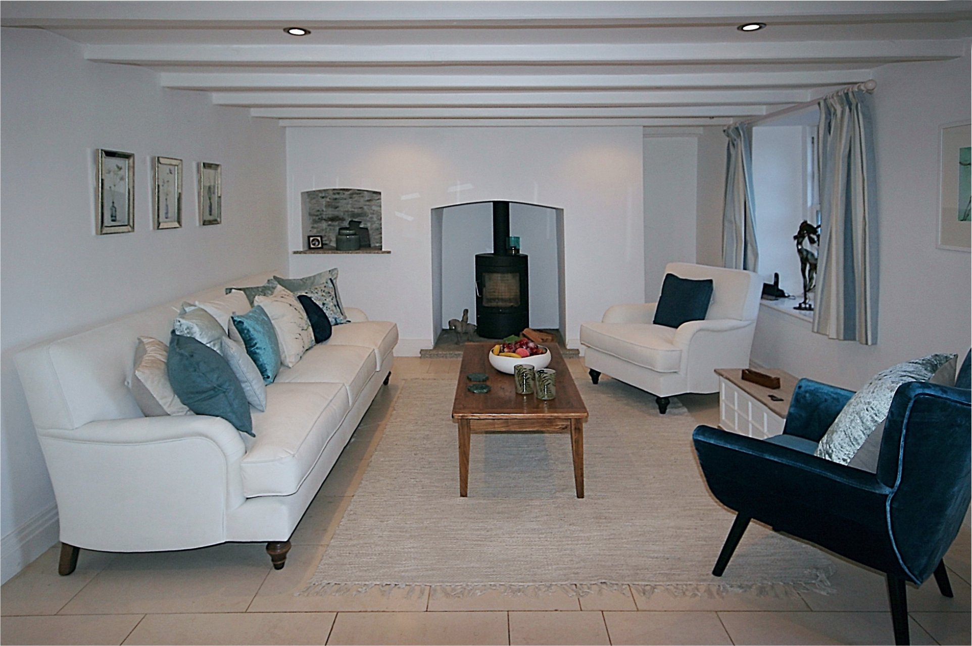 A wood burner is the focal point in this lounge which has white sofa and armchair and an alternate velvet blue chair.  There are blue and white cushions and an elm coffee table sits on a rug.