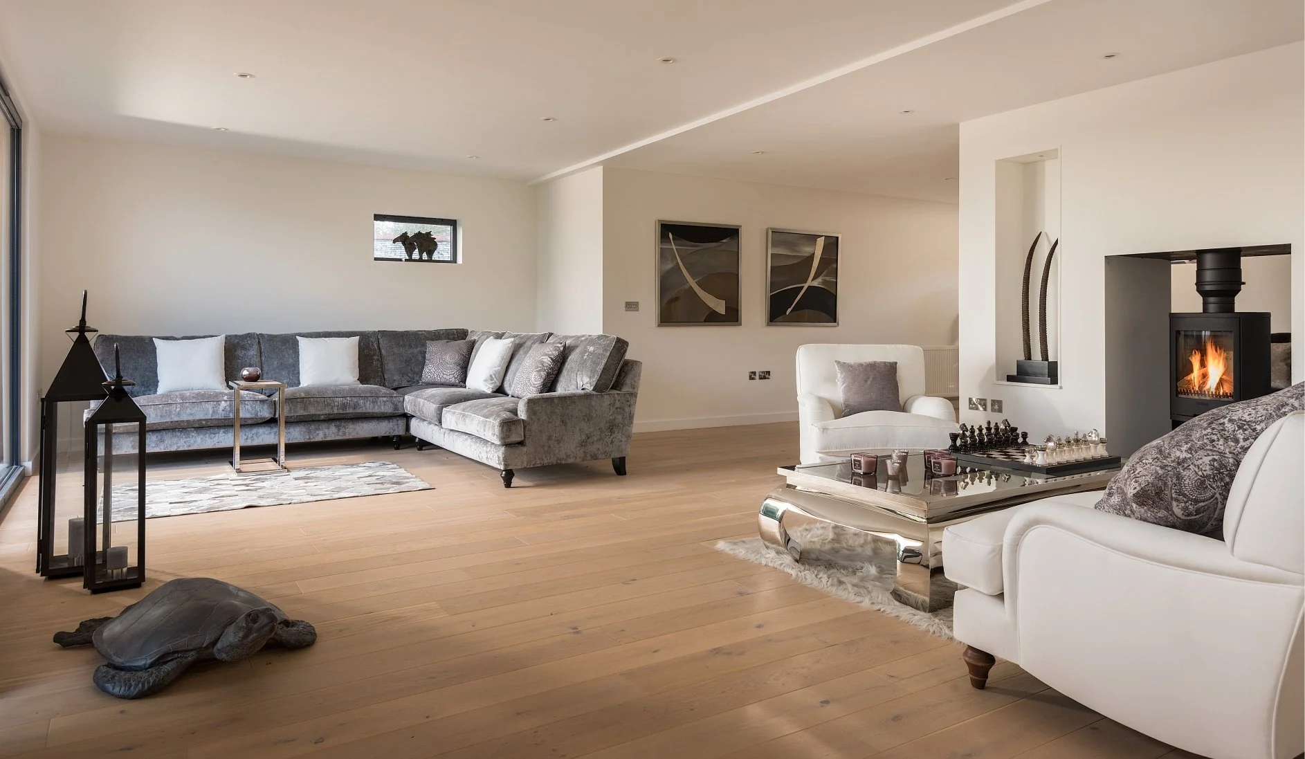 grey and white living room with oak floor.  There is a turtle decoration, pair of lanterns, horse ornament, chess set and striking wall art.  A wood burner is alight in the corner