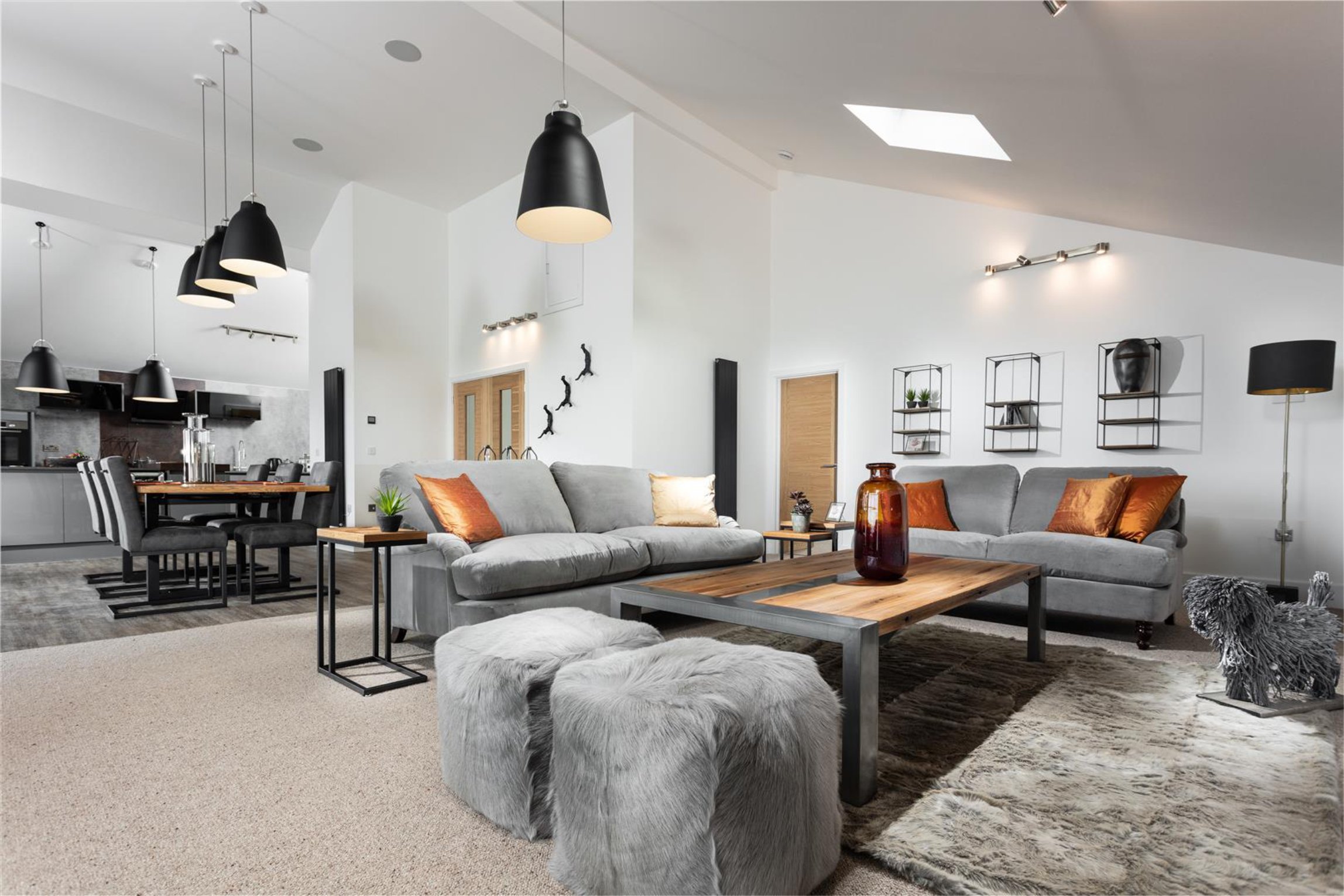 grey sofas and metal and wood coffee table have accents of burnt orange.  There is fluffy footstools and metal artwork as well as industrial lighting and black radiators. S bend dining chairs sit around an oak dining table