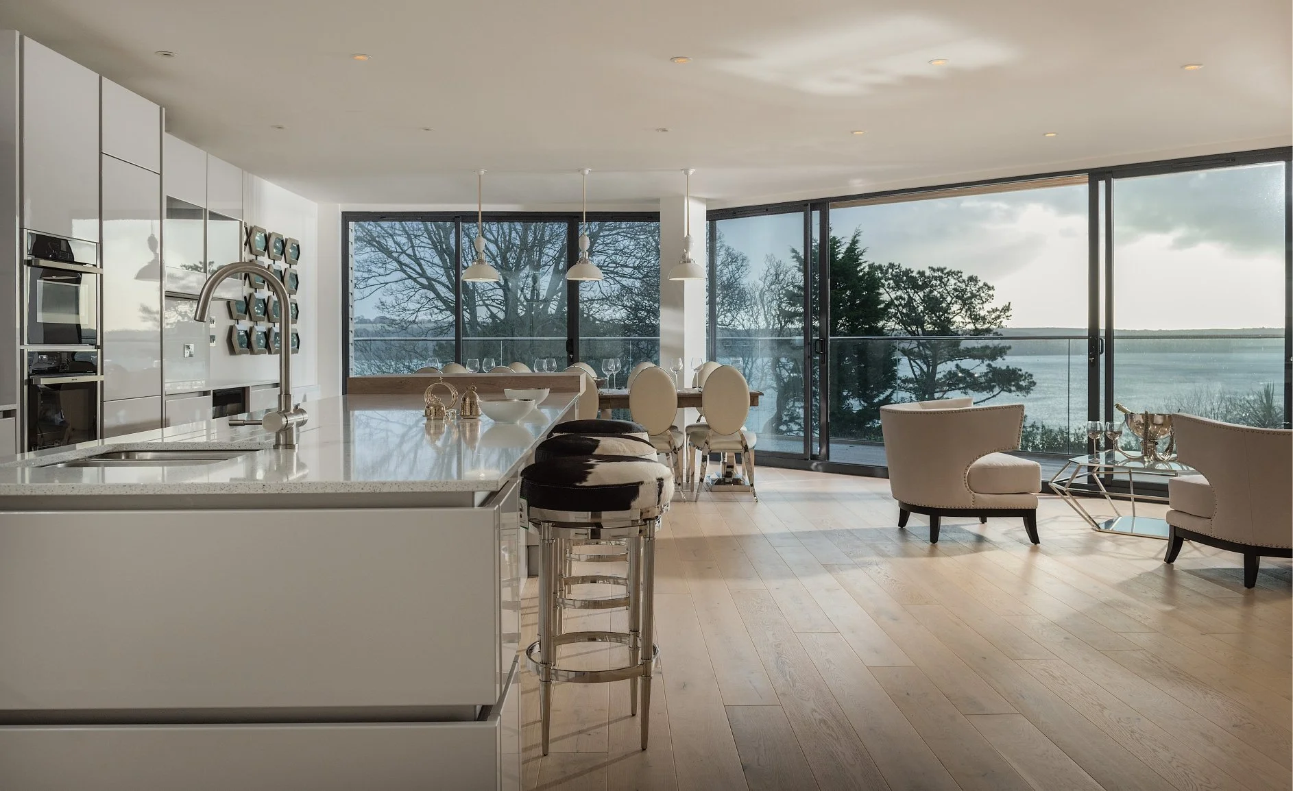 a white kitchen has massive windows overlooking trees and the sea.  There are cow hide bar stools and dining table with white dining chairs.  Casual seating with coffee table also feature