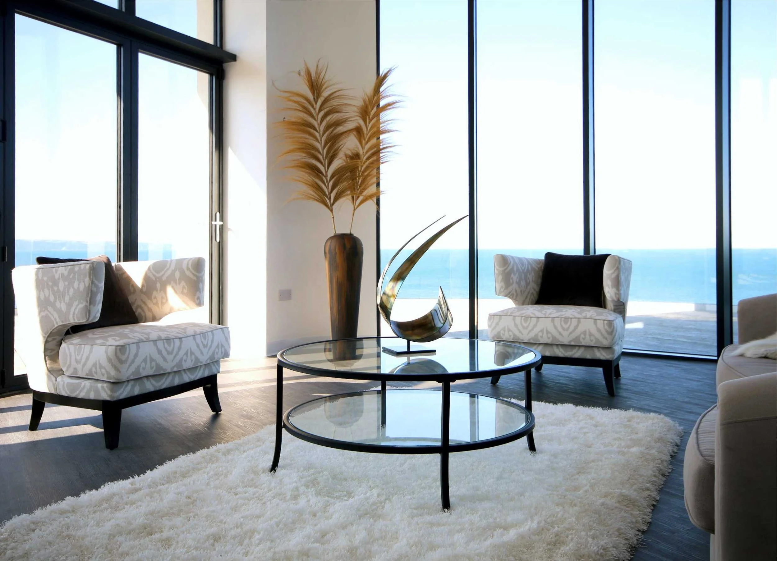 The lounge in this coastal house is themed in neutral tones to allow the sea views to be the focal point.  There are two chairs, a round glass coffee table, a curved metal sculpture and  a vase with tall grasses.  It was featured on Grand Designs