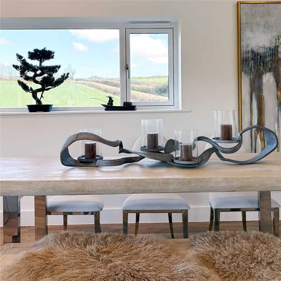 A deep solid wood dining table has stainless steel legs, on top is a large modern forged steel candle holder.  A bench has fur rugs and there are three white dining chairs.  On the window ledge behind there is a bonsai tree and a fisherman decoration