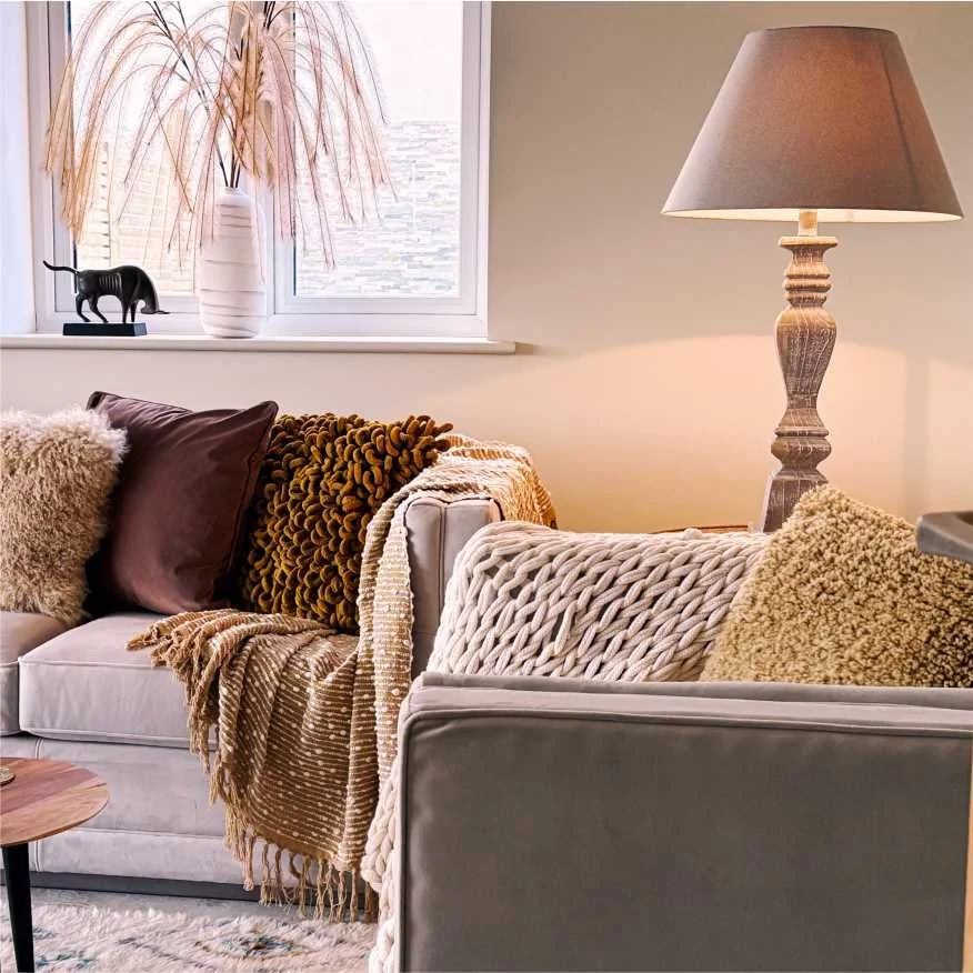 A hygge inspired living room with lots of textured throws and cushions in yellow, beige and ochre.  There is a tall wood table lamp, large grass decoration ad a stylish bull ornament