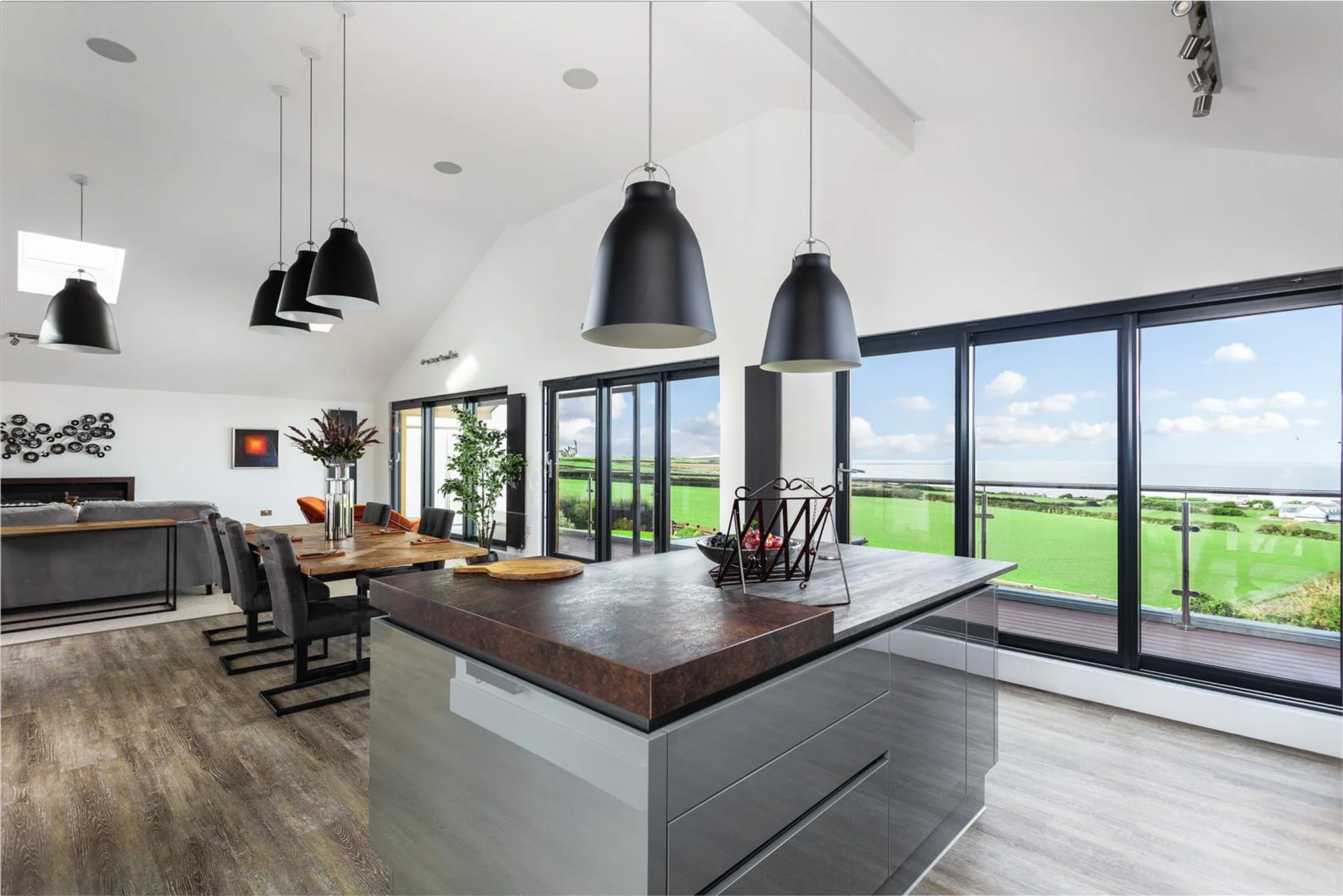 open plan living area with double height ceiling and drop industrial black lights.  There is metal artwork and foliage in the window.  An abstract orange and black framed art is on the back wall.