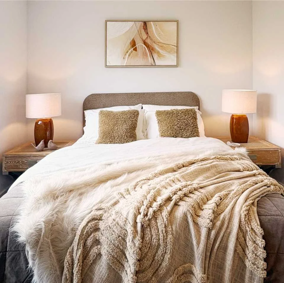 A casual yet sumptuous bedroom in taupe, browns and orange. Textured and fur throws sit atop a velvet quilted bedspread.  An abstract framed picture matches ceramic table lamps.  Two decorative birds are on the oak bedside table