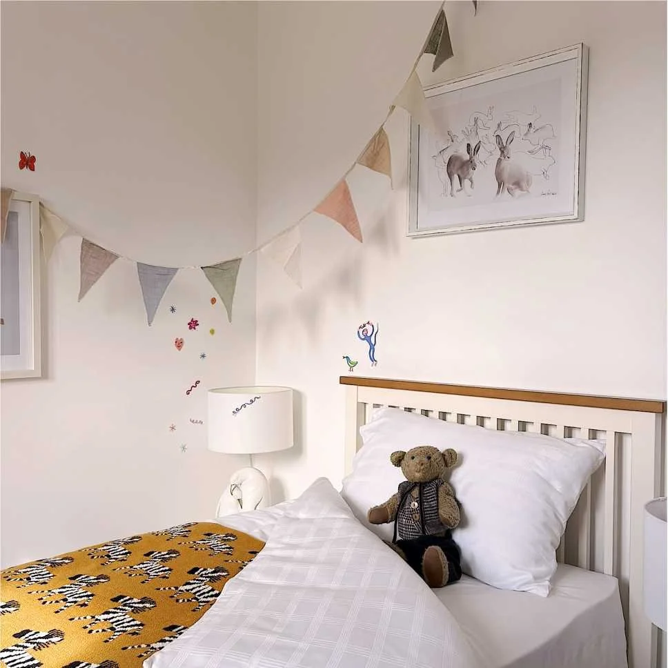 a banner in muted colours hangs above a bed decorated with a zebra mustard throw.  A teddy bear sits welcoming propped up on the pillow and a rabbit picture hangs above the white and oak headboard.  Playful stickers adorn the walls