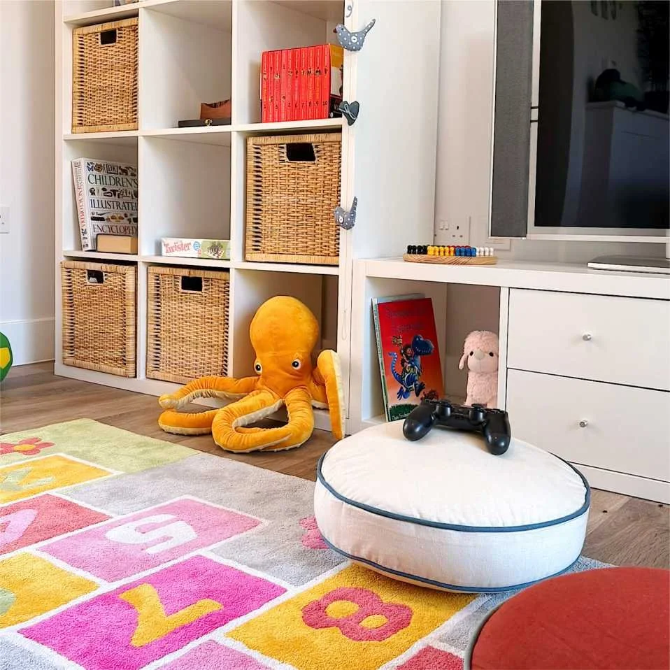 A colourful hopscotch rug is surrounded by a yellow octopus soft toy, floor cushions and play station.  A white shelving unit has wicker baskets, and toys, games and books