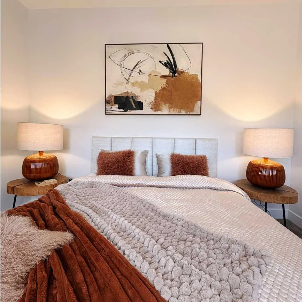 master bedroom with faux fur throws and fluffy cushions is set off by abstract wall art.  Two matching orange bedside lamps sit on natural wood tables