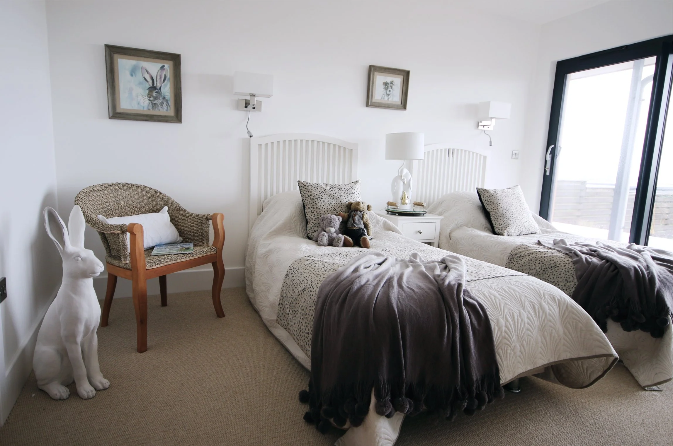 pictures of rabbit and dog are behind two single beds which have spotty cushions and grey throws with teddies and feature lighting.  A large stone rabbit is on the floor and there is a wicker chair