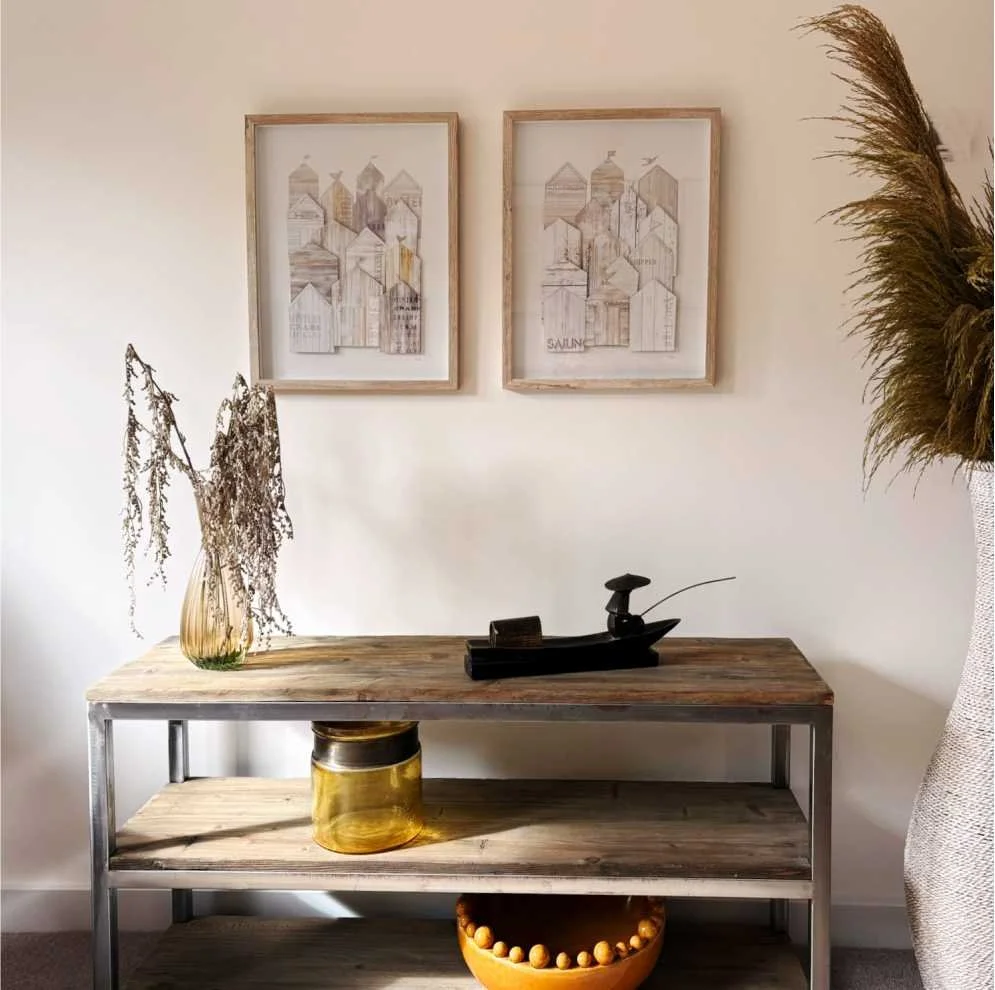 a casual lounge interior design idea with neutral and yellow colours.  A low wooden shelf has a vase of dried grasses, a fisherman decoration and yellow ball rimmed owl.  There is two coastal themed pictures on the wall