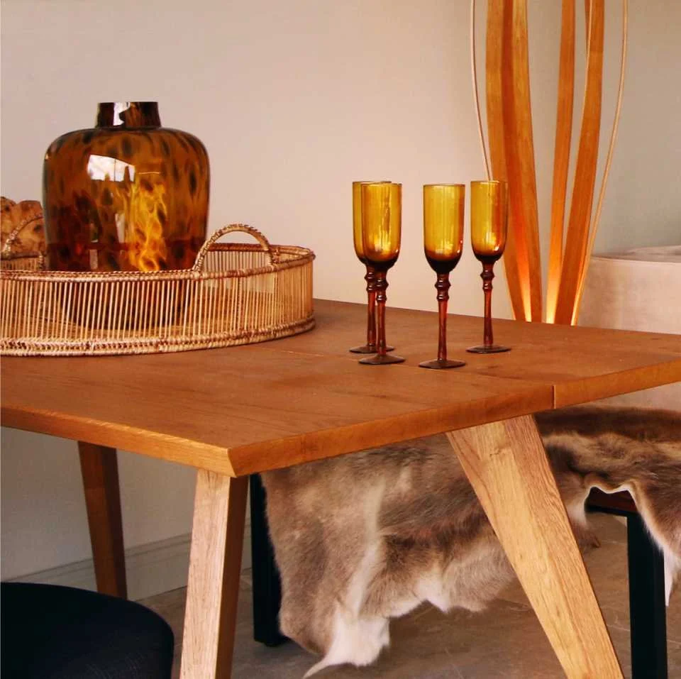 an oak dining table is decorated with yellow wine glasses, a wicker tray and a leopard print glass vase.  behind is a wood lamp and reindeer fur throw