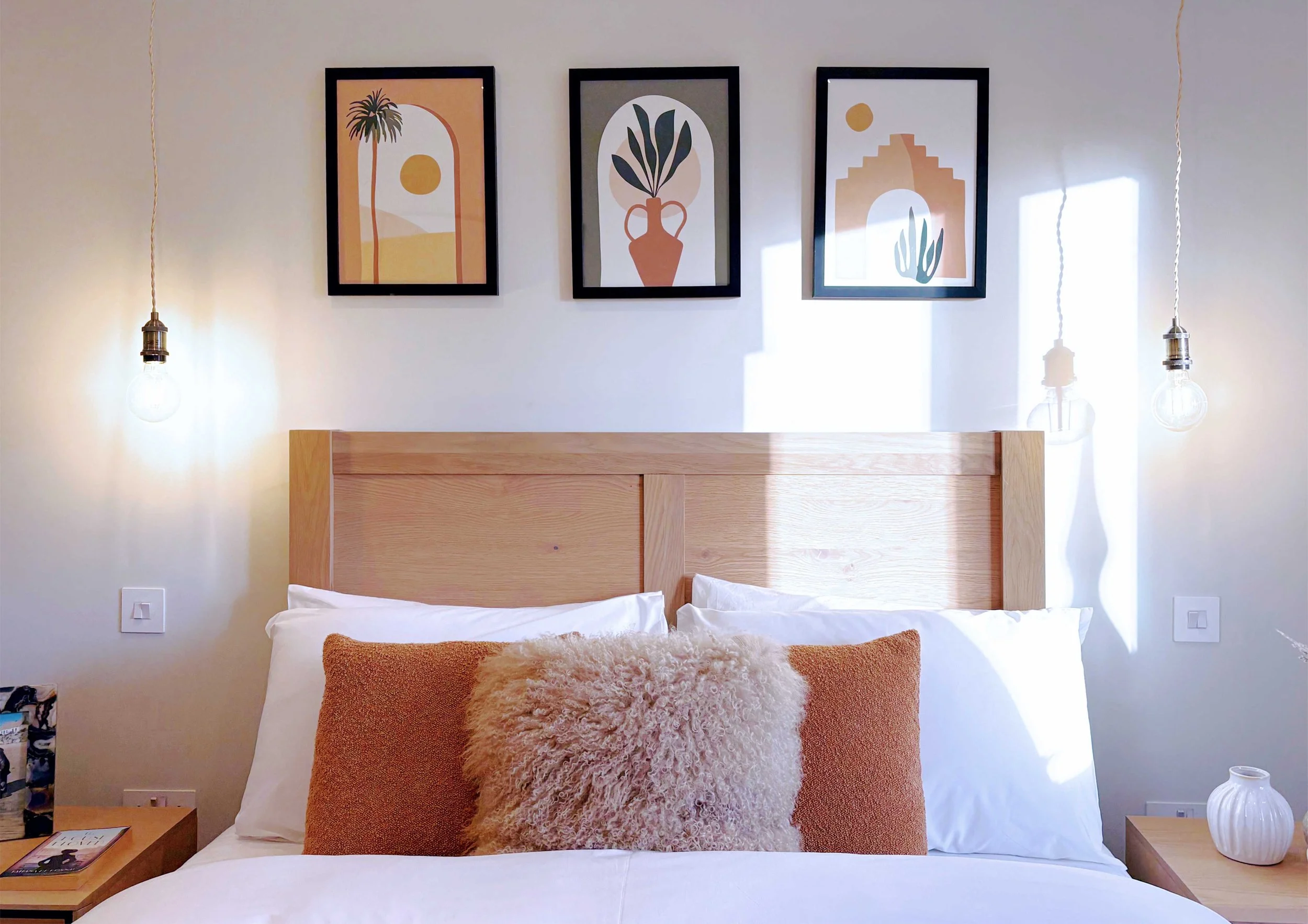 Wooden headboard and bedside tables with three  moroccan style graphic framed prints.  Fluffy and textured brown cushions set against plain white bedding