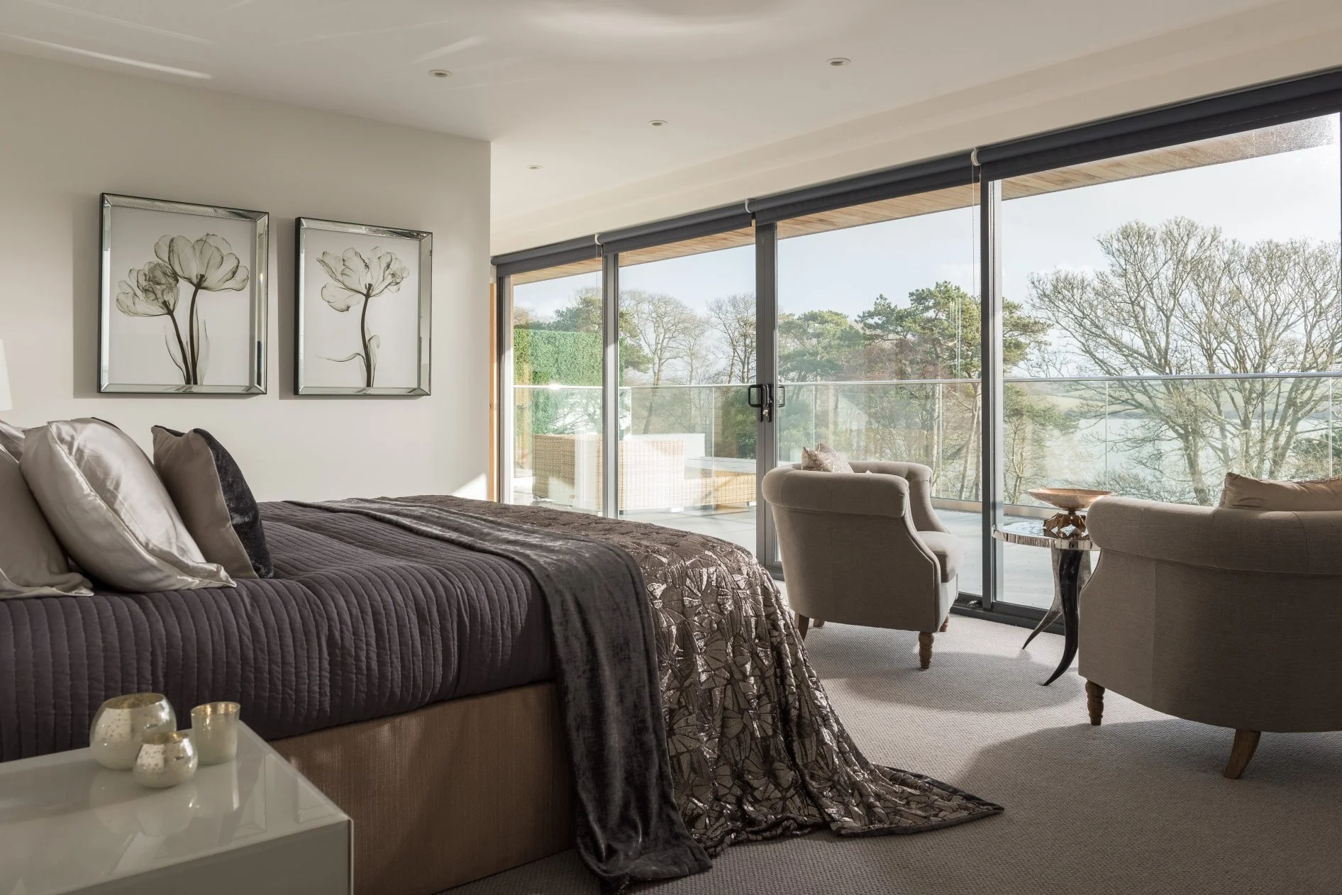 Bed with draping bed throws, large windows opening onto a balcony and two easy chairs with coffee table sit in the window.  Two xray pictures of flowers are on the wall