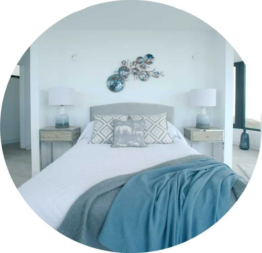 A modern bedroom with a neatly made bed, decorative pillows, and a grey blanket. There are two matching nightstands with white lamps and a blue vase on the right. A piece of wall art depicting blue circles and a branching design decorates the white wall behind the bed. A large window on the right lets in natural light.