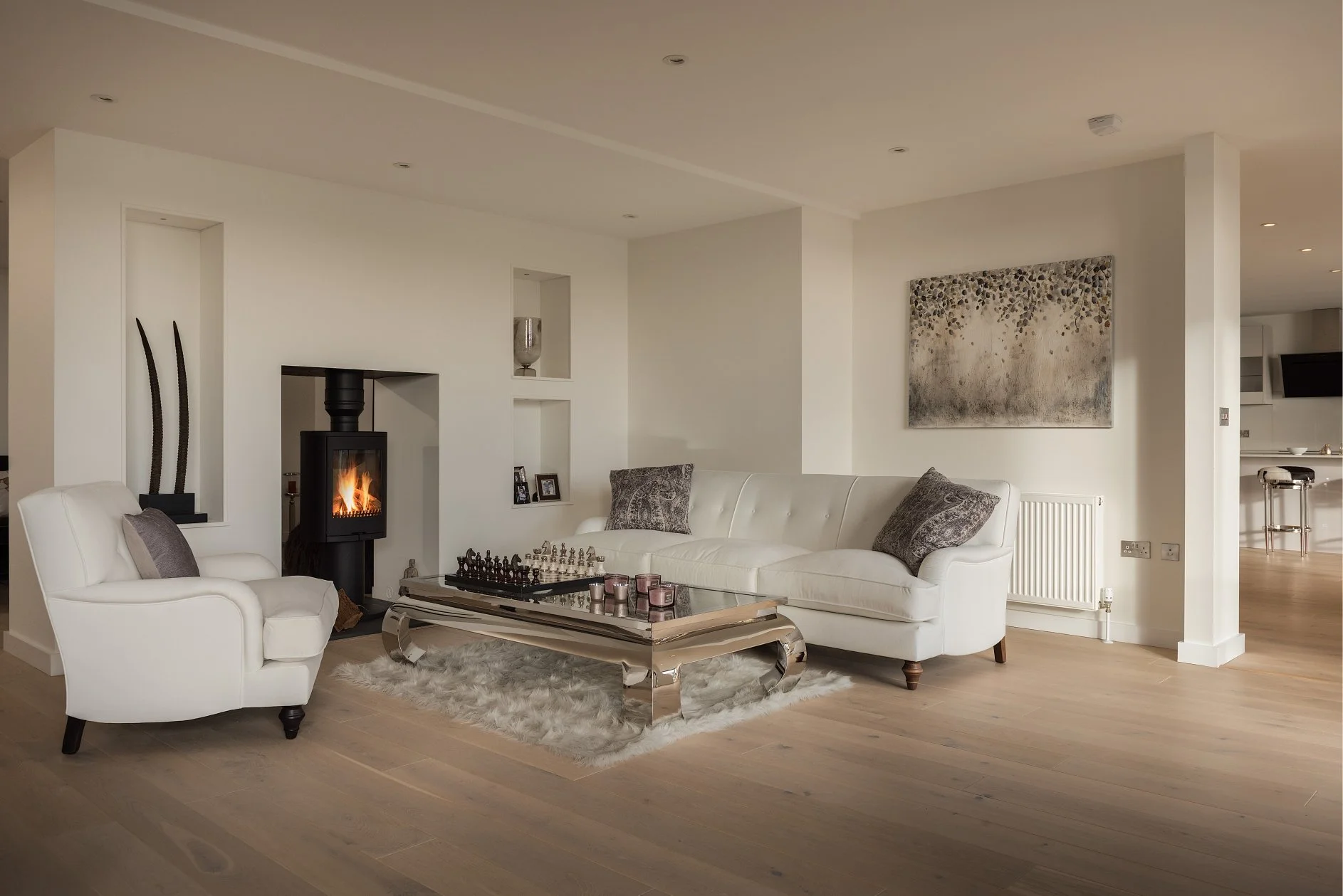 white walls and white sofa and chair with grey artwork and cushions .  A chess set sits on a chrome coffee table.  There is a woodburner, grey accents and a fluffy rug