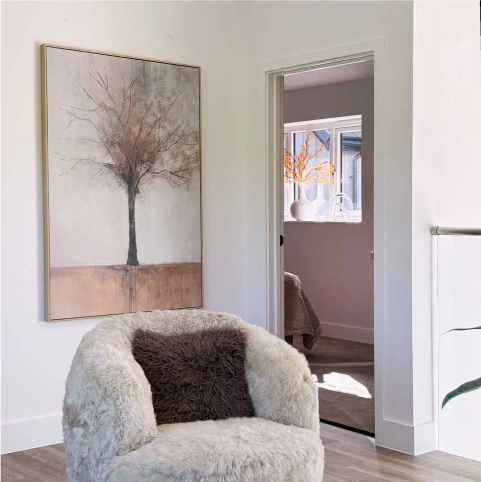 A sheepskin round chair has a brown fur cushion.  Behind is a large framed print of a stylised tree in coppery orange tones and brown.  Through a doorway a vase of orange stems can be seen echoing the shape of the tree