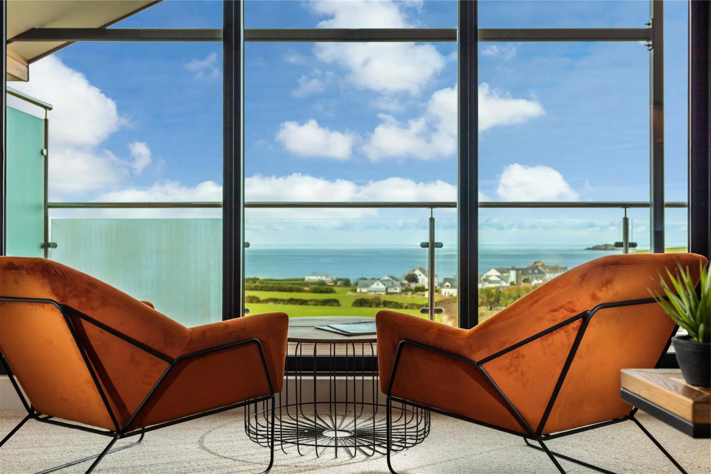 two modern pumpkin orange chars look out across far reaching views.  There is a cage style coffee table with magazines on it.