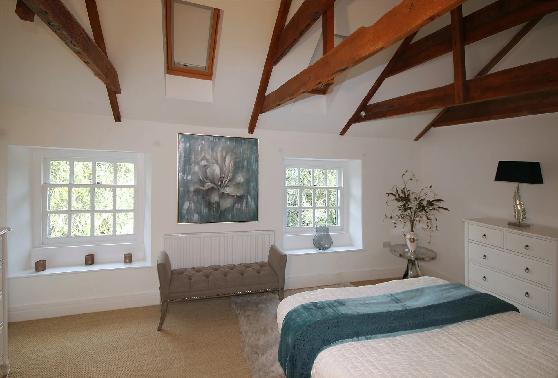 blue and white bedroom with rural aspect and vaulted ceiling with wood beams.  There is an ottoman stool and an oil painting of a flower.  A blue throw is on a white bedspread on the bed