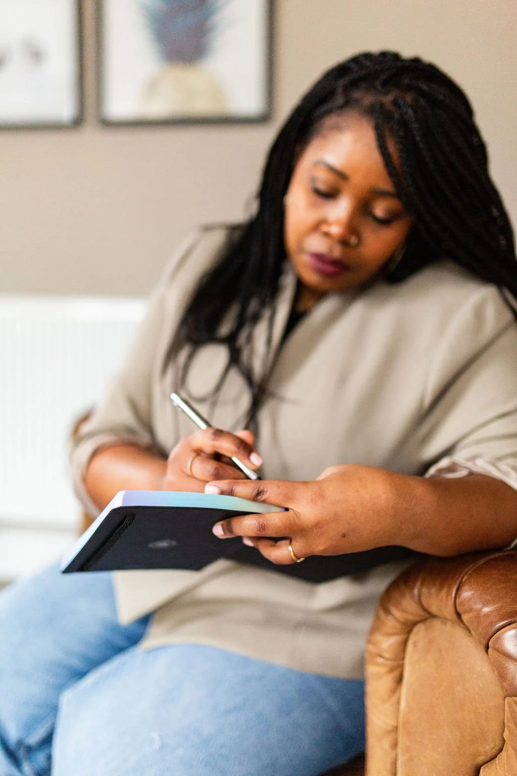 A woman with dark braided hair sitting on a brown leather couch, writing in a black notebook with a silver pen, in a cozy room with framed pictures on the beige wall behind her.
