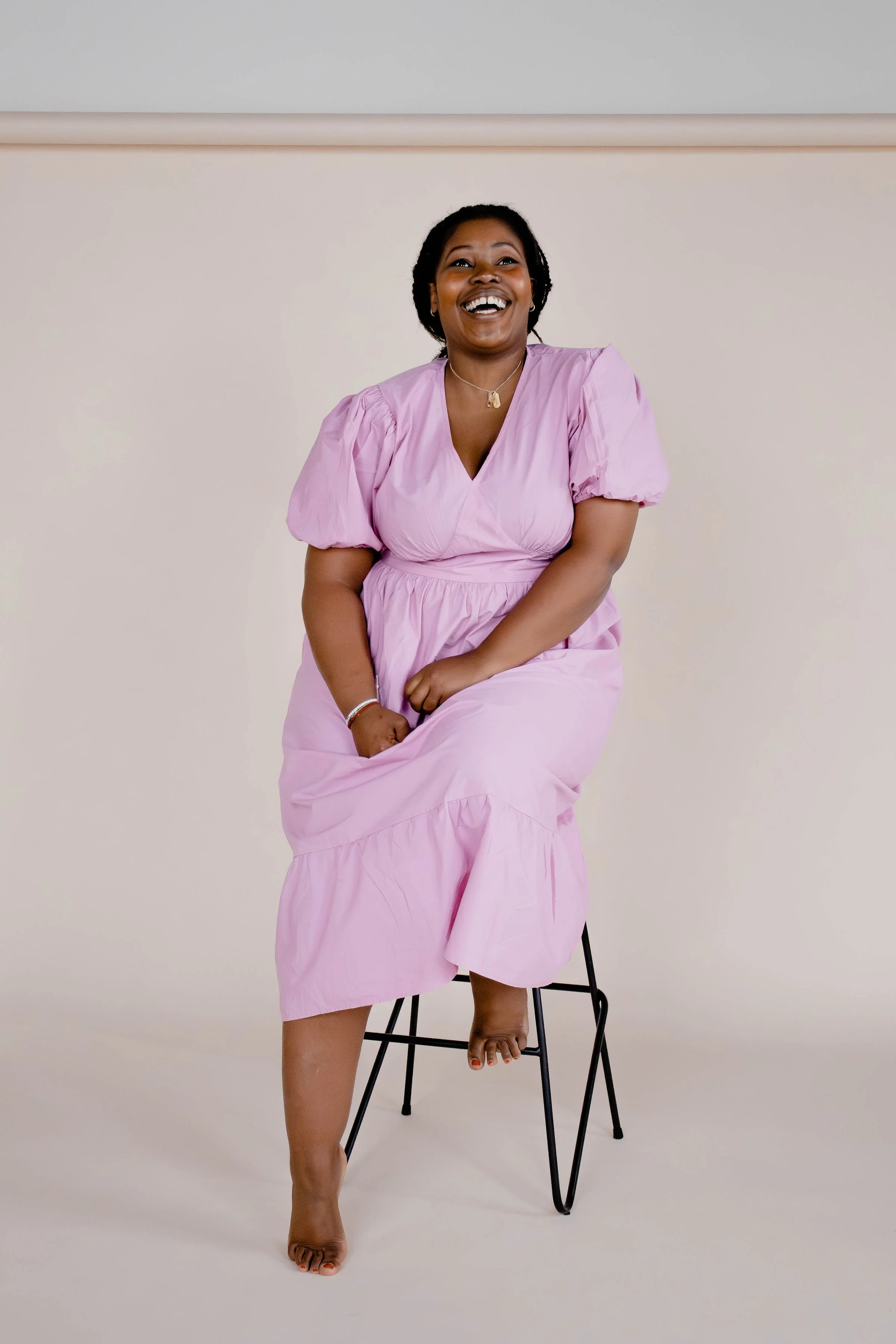Smiling person in a pink dress sitting on a stool against a neutral background.