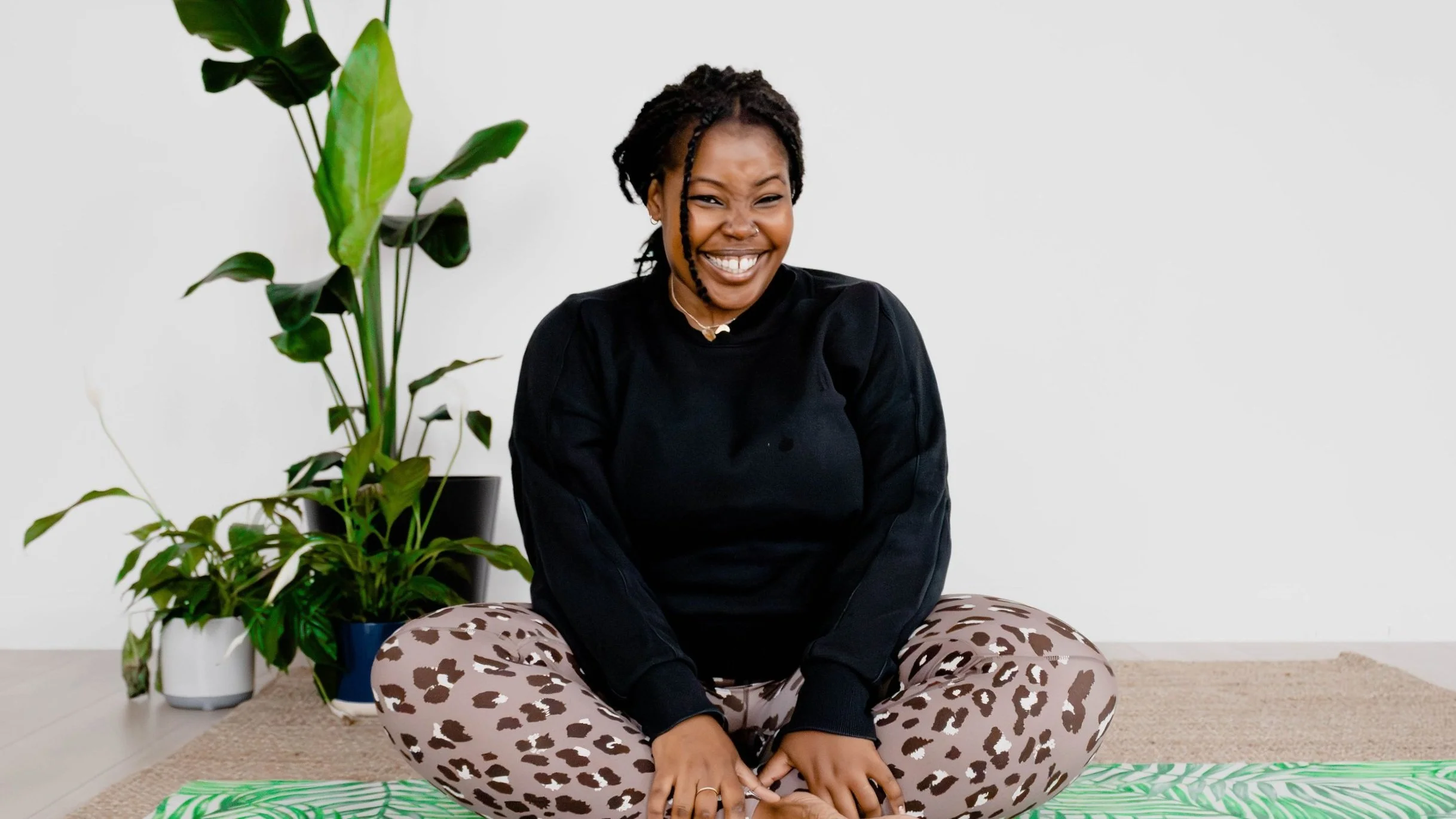 Person sitting cross-legged on the floor, smiling, wearing a black sweater and leopard print leggings, with green plants in the background.
