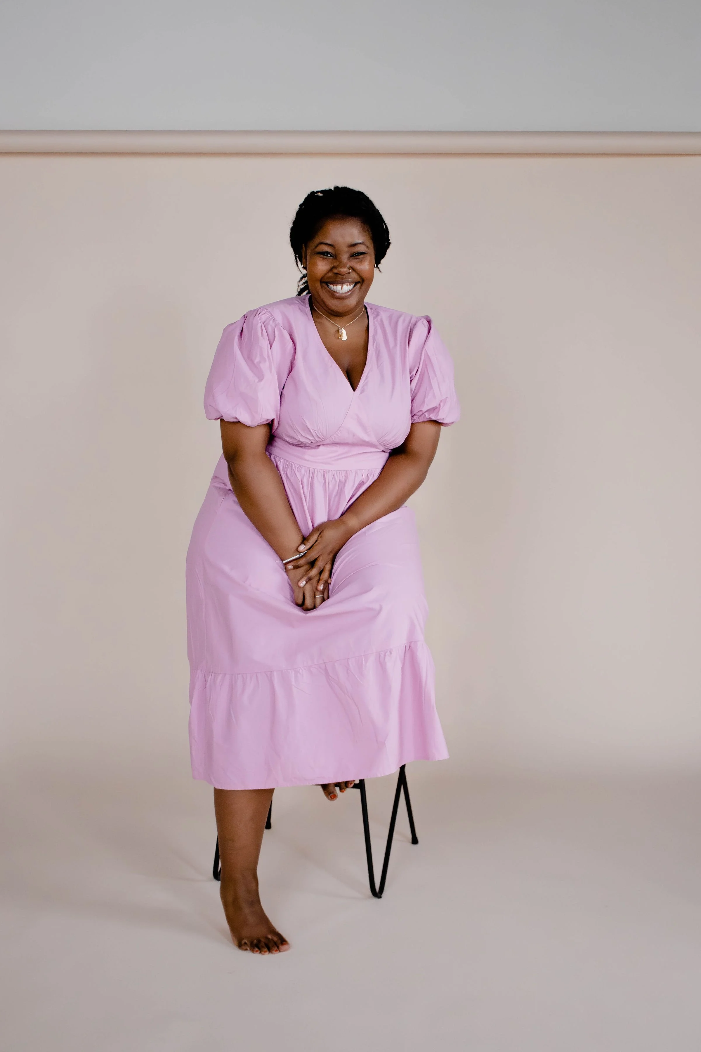 Smiling woman seated in a light pink dress, barefoot, on a stool, with a neutral background.