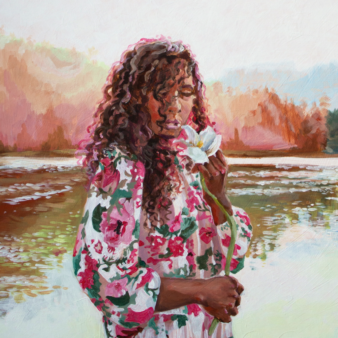 A woman with dark skin and curly hair holding a white lily near her face by a body of water with a colorful forest in the background.