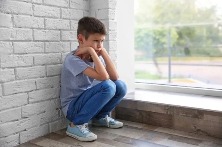 Young boy brown hair with blue jeans and blue tee shirt crouching in a corner by window and white brick wall