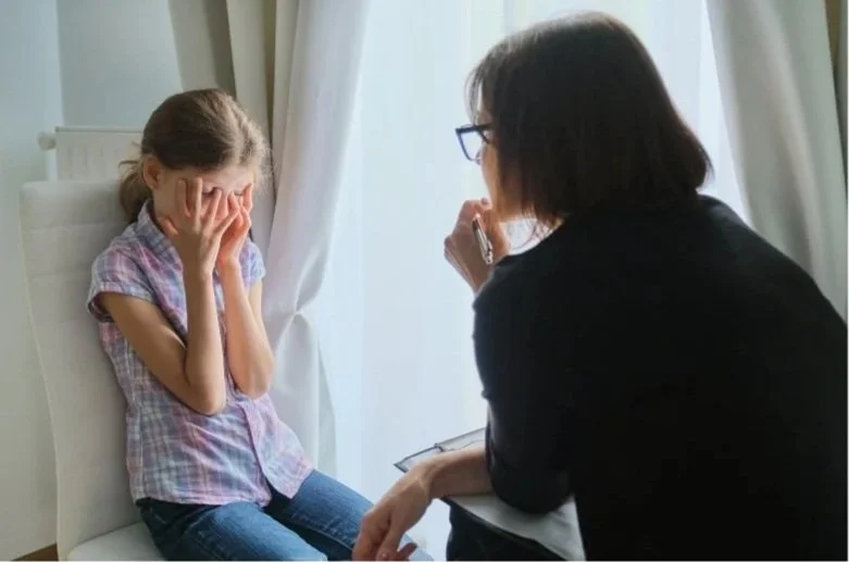 Young girl sitting with her hands on her face in front of a woman with dark hair and glasses  leading forward