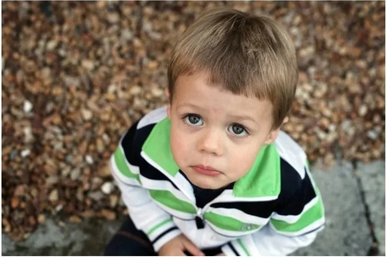 Young boy with green, black and white striped shirt looking up at the camera with a sad face