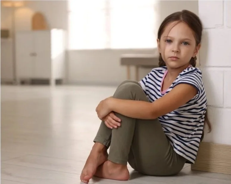Young girl in striped shirt and olive pants sitting on the floor against a white wall