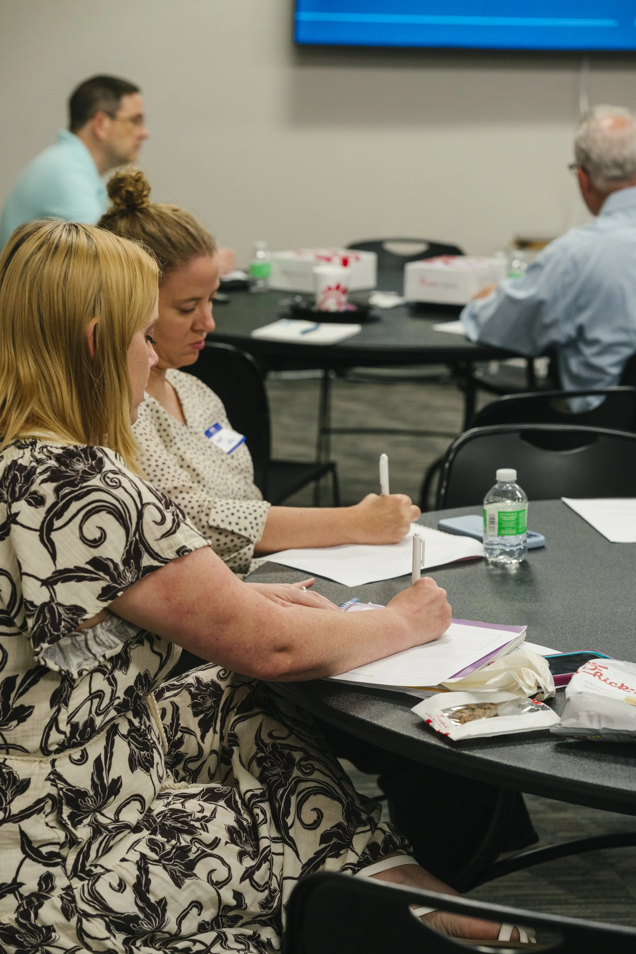 People sitting at a conference table taking notes, with food and drinks, in a professional setting.