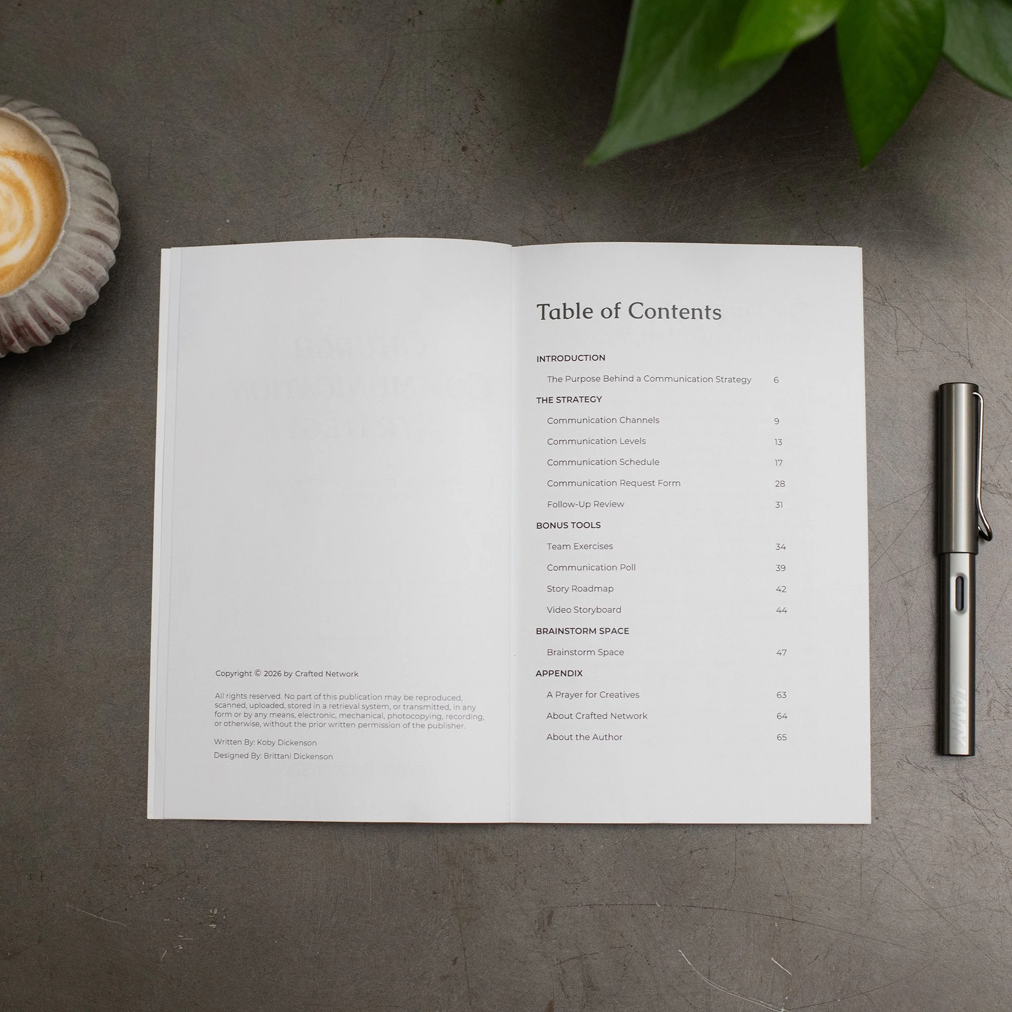 A photograph of a table of contents page from a book, placed on a dark gray surface. A green leafy plant is partially visible at the top right corner, a ceramic cup with latte art on the left, and a silver pen on the right.