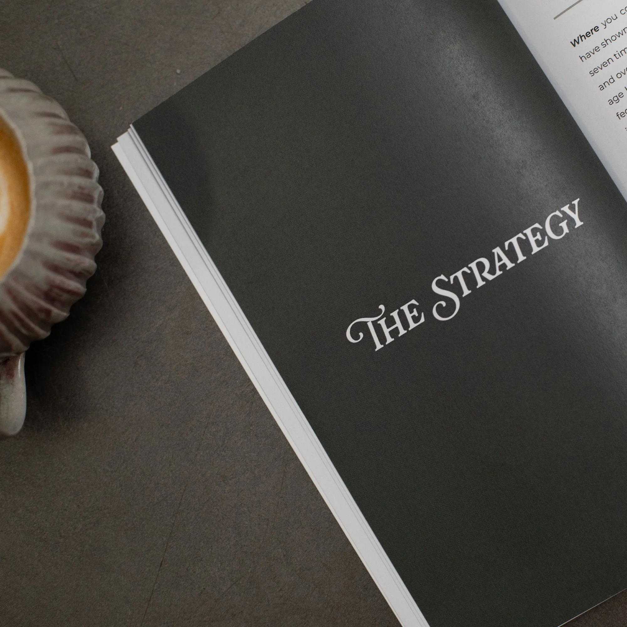 Close-up of a rolled muffin beside a black and white book titled 'The Strategy'. Part of a white sheet of paper is visible in the top right corner.