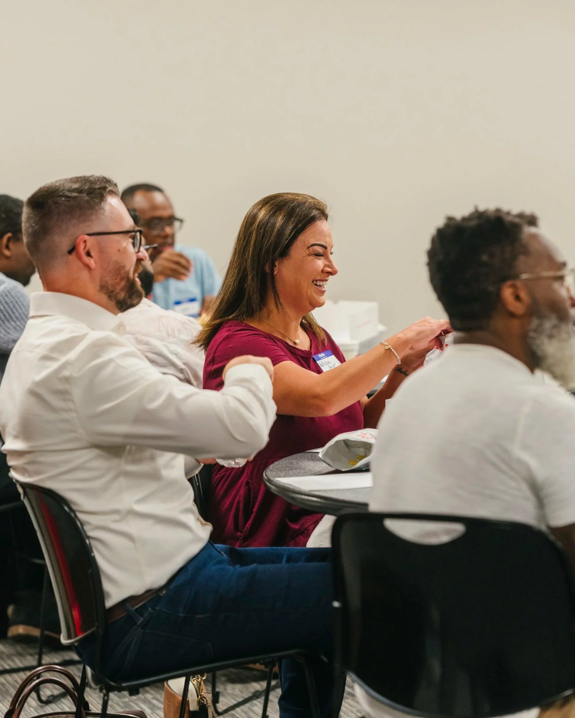 Group of diverse individuals in a conference room, sitting at tables, smiling, engaging, and listening attentively during a professional seminar or workshop.