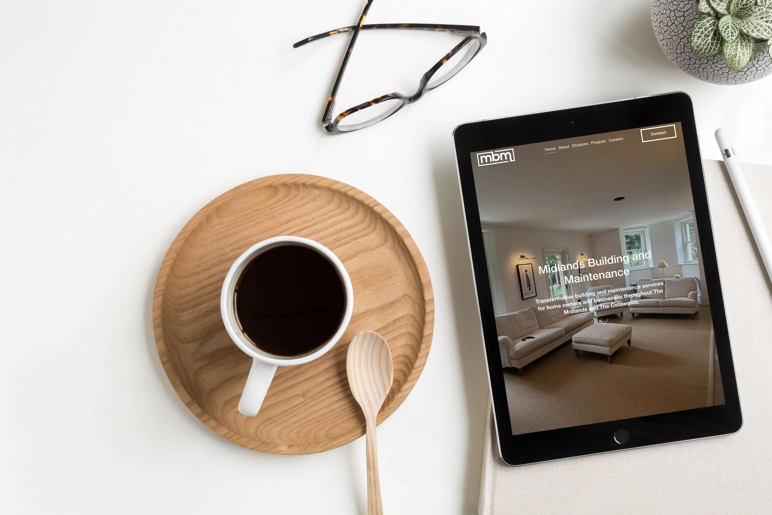 Flat lay of a smartphone displaying a website, a cup of coffee on a wooden charger plate, a pair of glasses, and two wooden spoons, set on a white table with a potted plant.