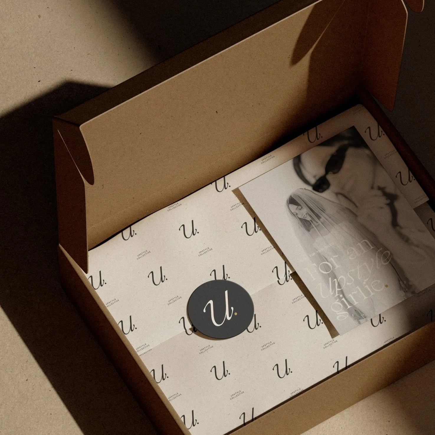 A cardboard box with 'U.' printed repeatedly inside it, containing a booklet titled 'Rising Up & Girlie' and a round black sticker with a white cursive 'U.' on it.