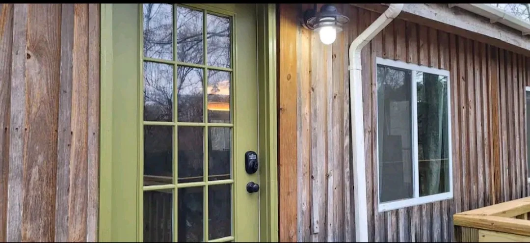 Close-up of a wooden house exterior featuring a green door with glass panes, a modern black door lock, a window with a white frame, a black outdoor light fixture, and a white gutter pipe.