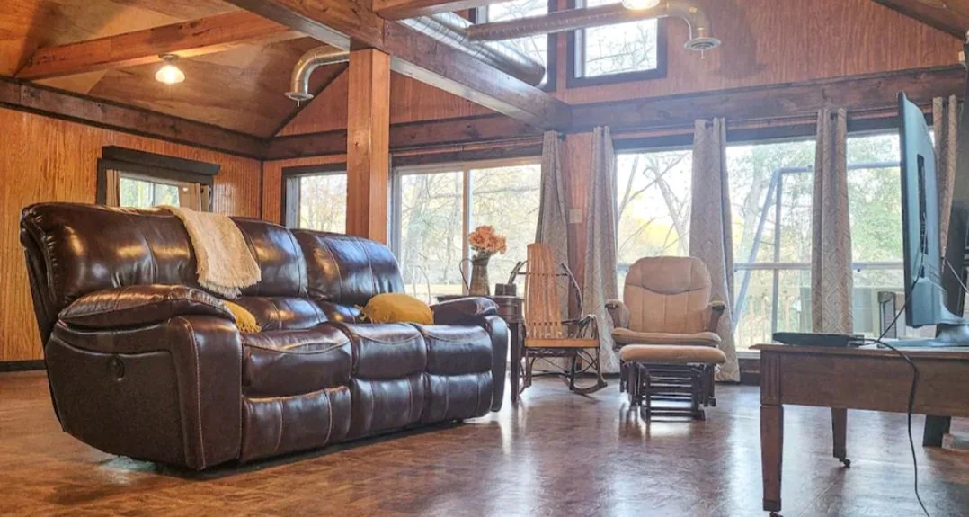 Living room with leather sofa, armchair, rocking chair, TV, large windows with curtains, wood beams, and wooden walls.