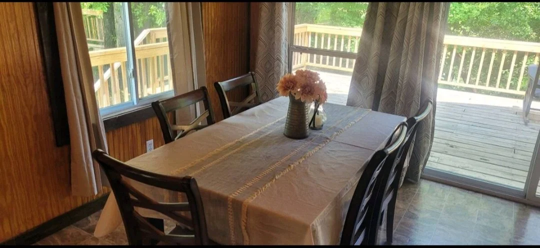 Dining table with a beige tablecloth, a vase with pink flowers, set in a room with wood paneling and large windows with curtains, overlooking an outdoor wooden deck with a wooded area in the background.